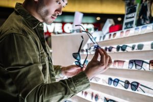 Side view of crop bearded Asian male customer picking sunglasses while standing near shelves in modern store