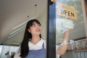Young female entrepreneur smiling while hanging open sign on glass door, representing small business, startup, and welcoming customers at local shop or cafe.